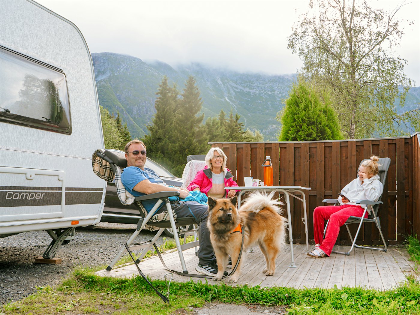 Family with dog sits outside caravan and laughs. Photo