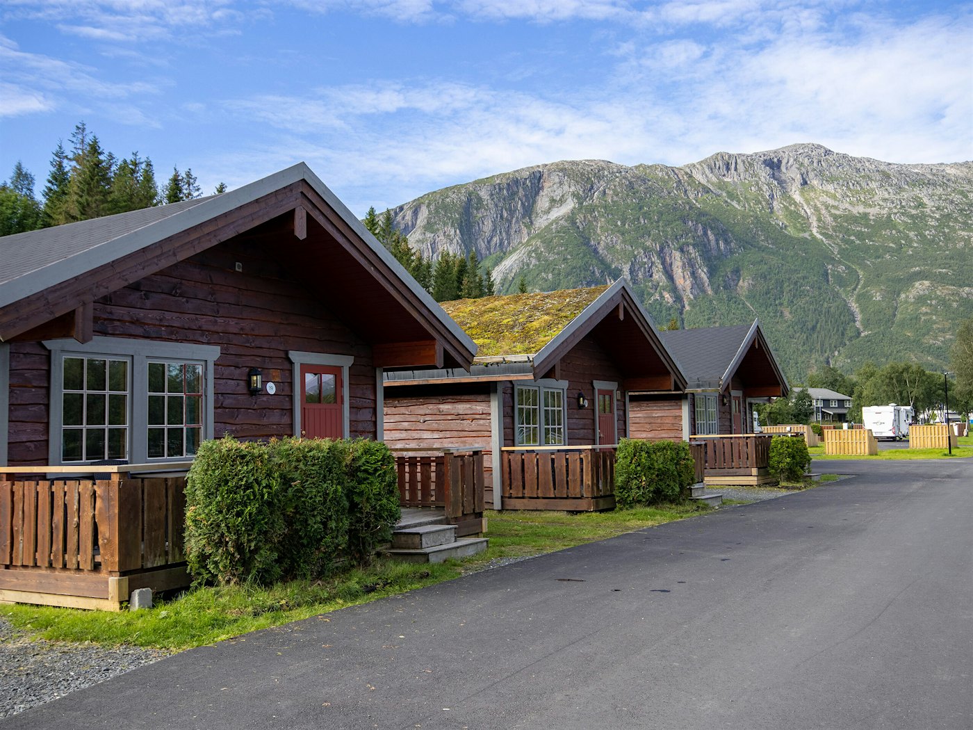 Three log cabins with grass on the roof and green lawn around, with mountains in the background. Photo