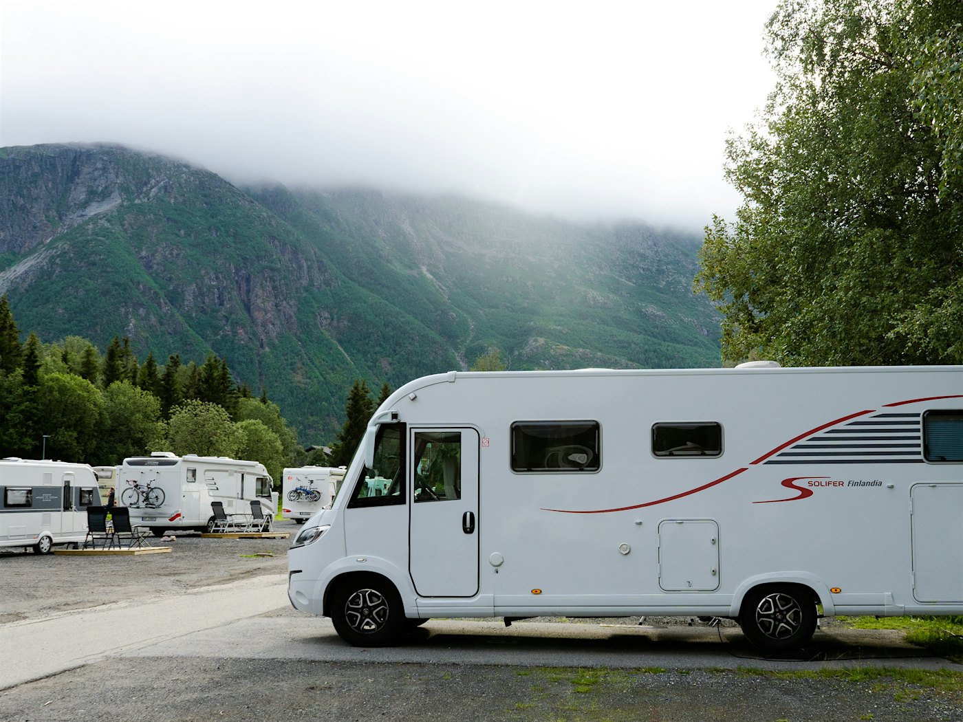 Bobiler og campingvogner står på campingplass, med høye fjell i bakgrunnen. Foto