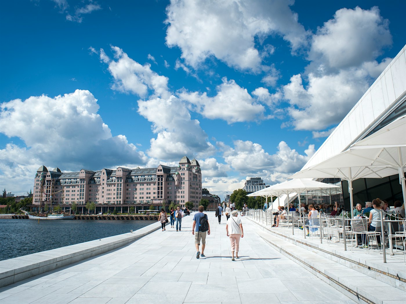 Passage in white marble with the opera house and restaurant on the right and the Oslofjord on the left. People walk in the passage. In the background you can see the city of Oslo.