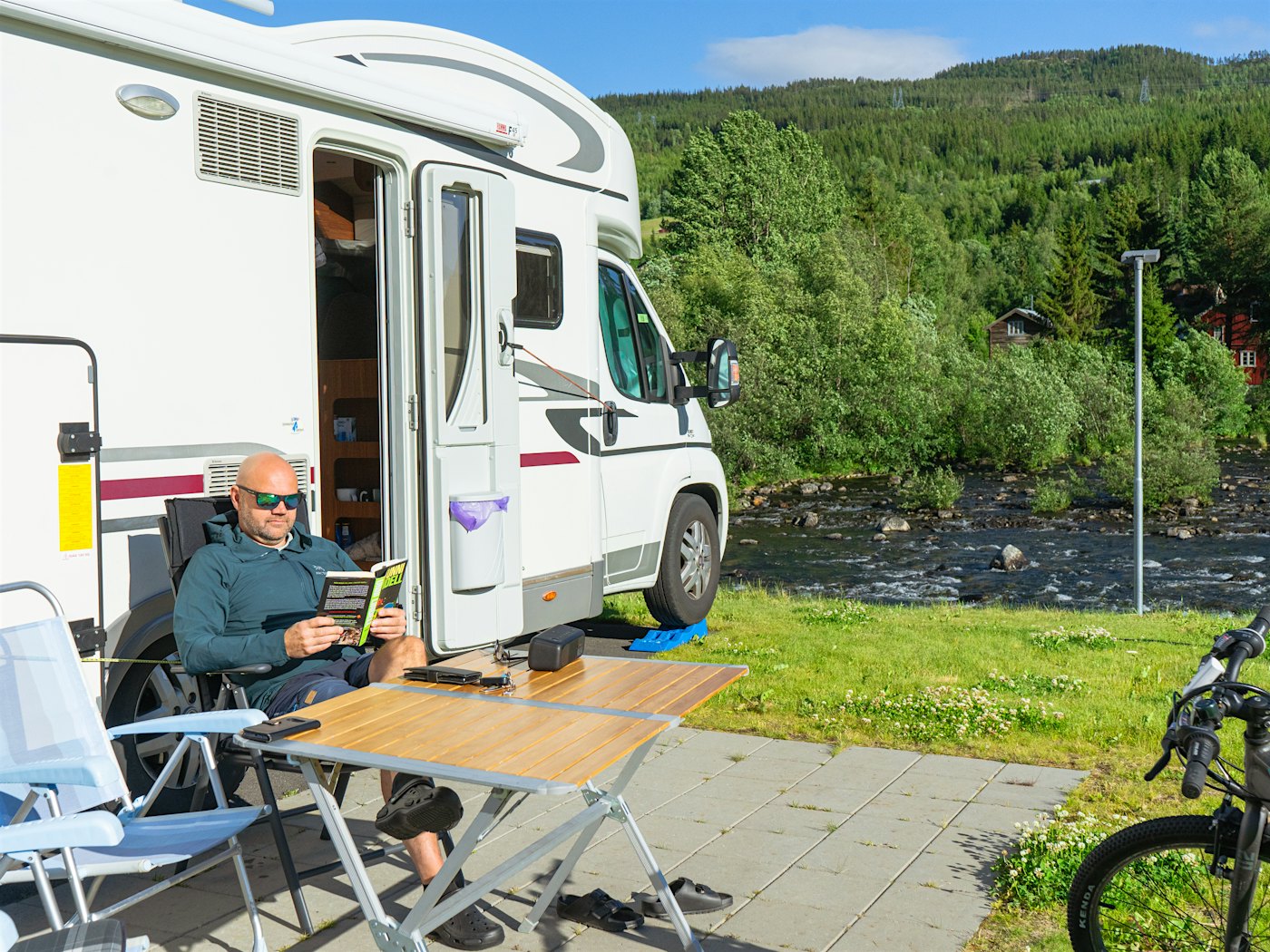 Man sits outside motorhome reading a book. River in the background. Photo