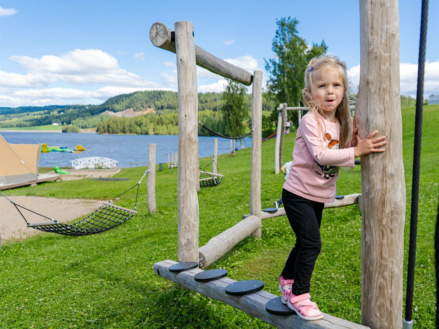 Jente på lekestativ på lekeplass i tre, med Mjøsa i bakgrunnen. Foto