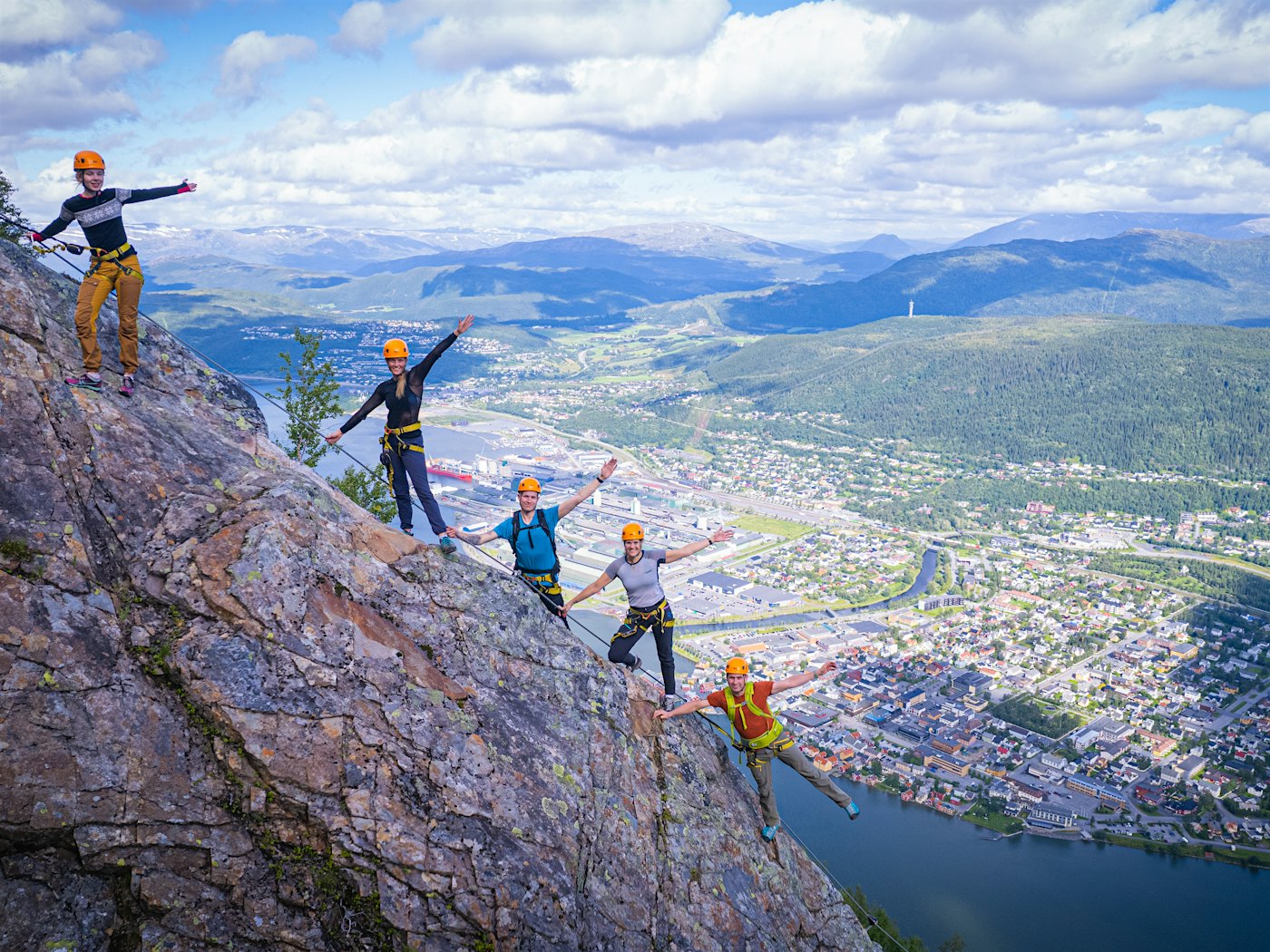 Fem personer strekker armene til værs mens de står i fjellside på vIA Ferrata-løype. Mosjøen og fjell i bakgrunnen. Foto