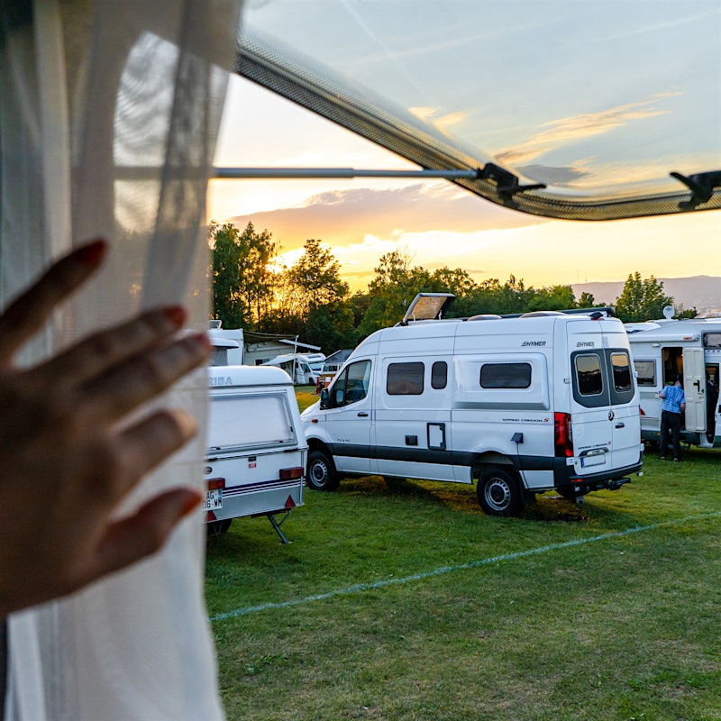 ånd skyver tynn gardin til siden i campingvinduet. Man ser utover campingbiler på gresslette i solnedgang.