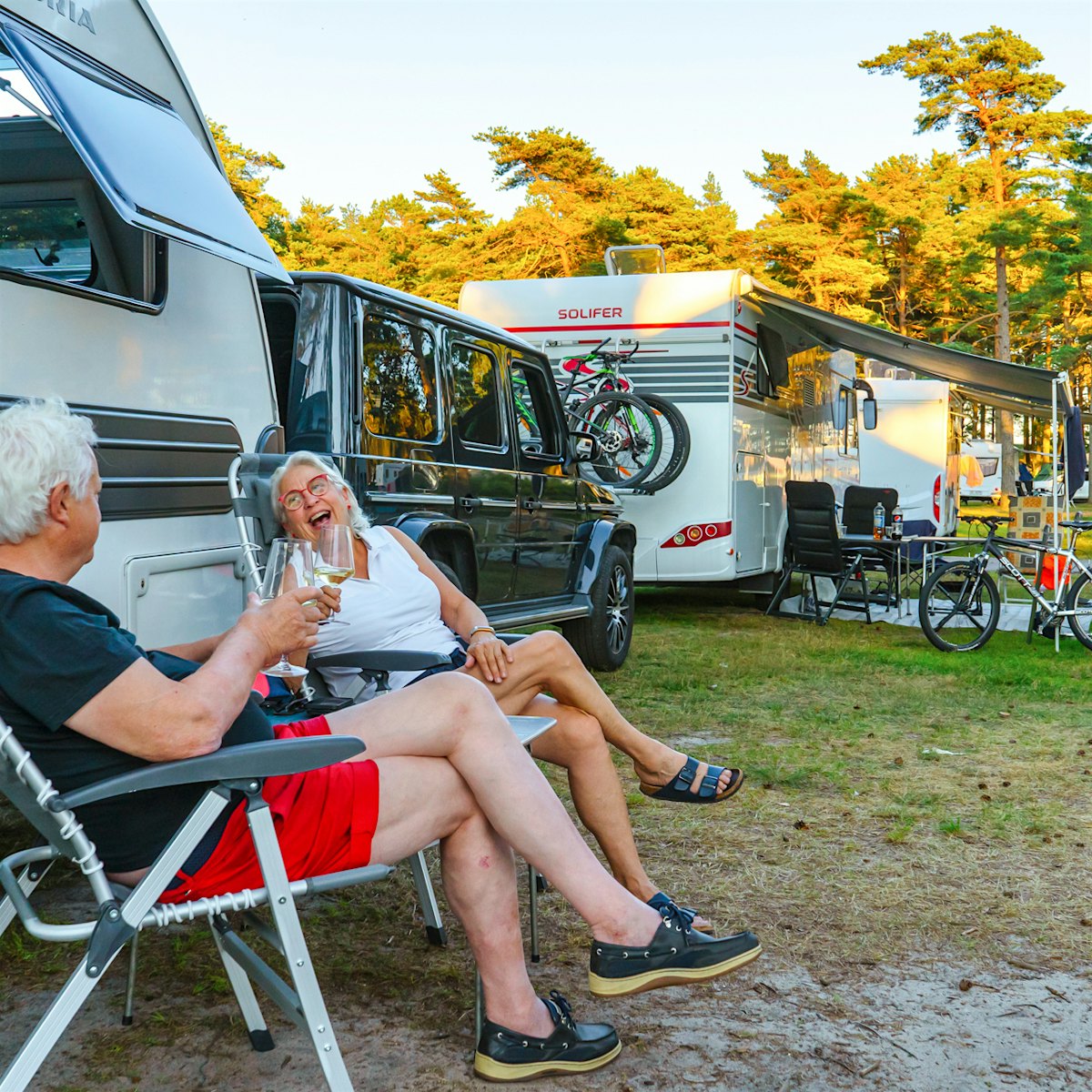 A married couple sits in front of their caravan, laughing and toasting two glasses together in the evening sun. Photo