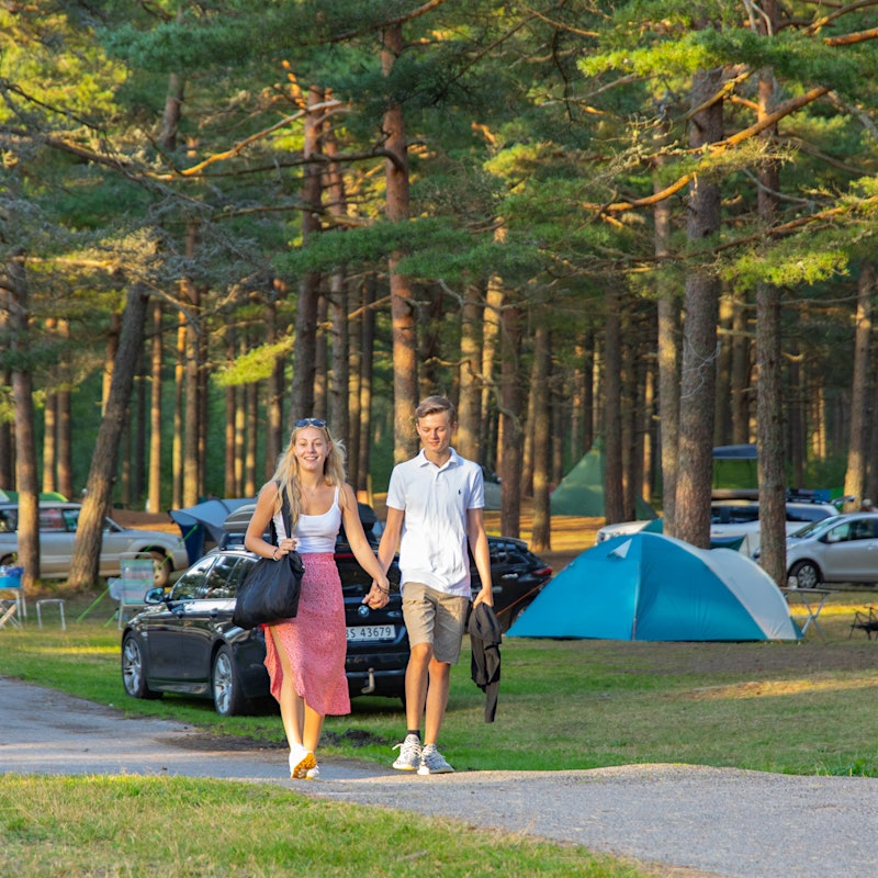 Ung kvinne og mann holder hender langs stien på campingplass. Kvinnen har veske under armen. Rundt dem er det trær, biler og telt.