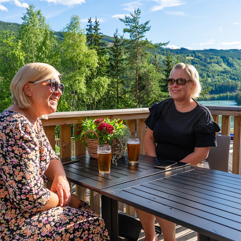 Two ladies are sitting at an outdoor terrace, smiling and enjoying themselves. View to trees, mountains and water. Photo