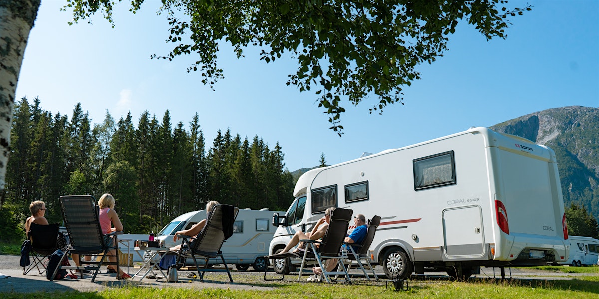 A group sits outside in the fine weather with their motorhomes on their sides. Mountains and trees in the background. Photo