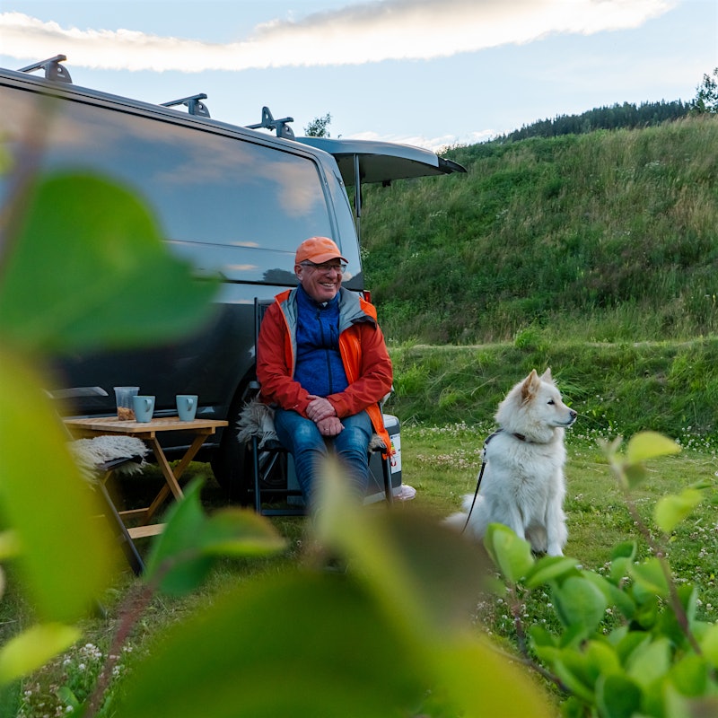 En smilende mann og hvit hund sitter foran en svart bobil. Det er busker i forgrunnen og grønn ås i bakgrunnen