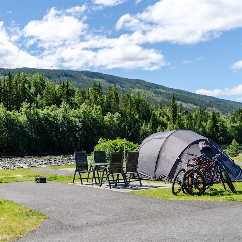 Telt står på teltplass med fire stoler og sykler foran. Utsikt til elv, skog og fjell.