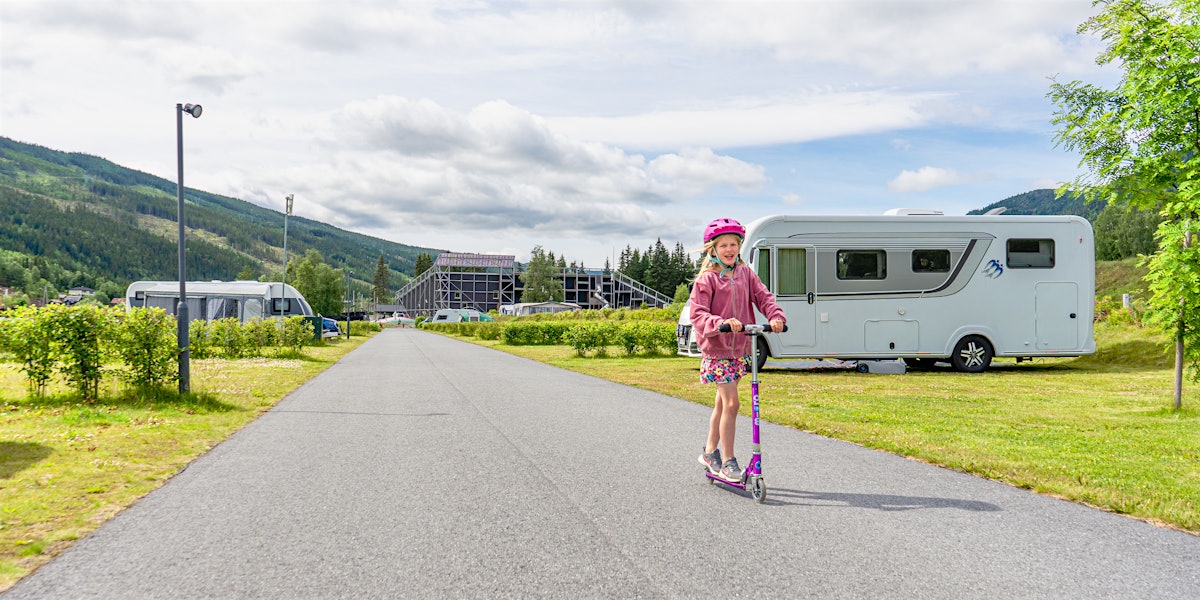 Mädchen steht auf einem Roller auf einem Campingplatz mit Spielplatz und Bergen im Hintergrund. Foto