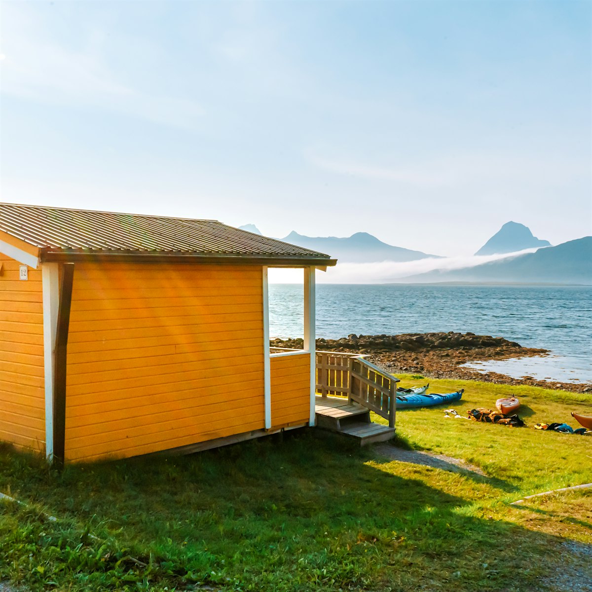 Gul hytte med utsikt til hav og fjell, med kajakker liggende foran hytte.
