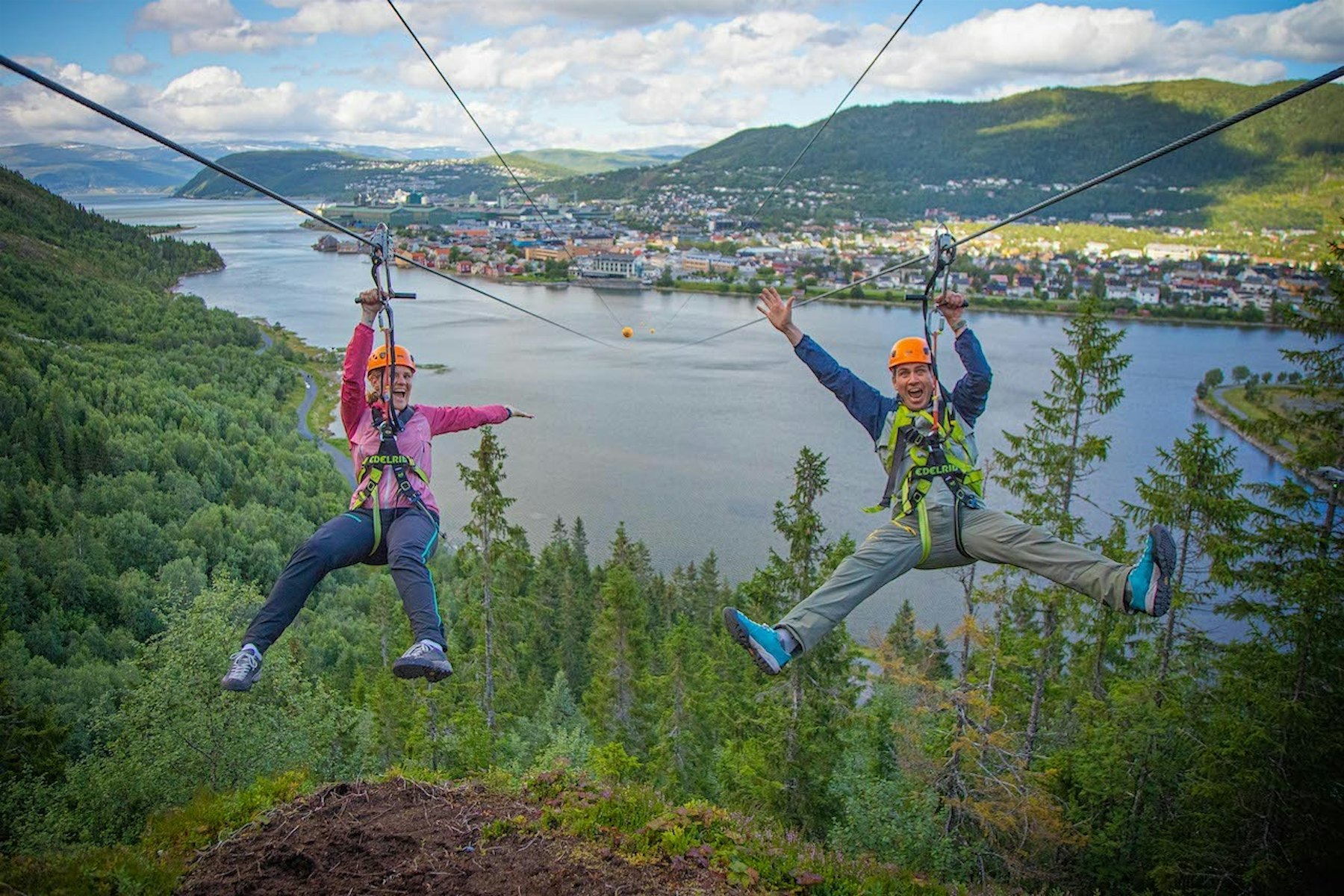 A man and a woman are hanging on a zipline and smiling big with their arms in the air, with Mosjøen as a view in the background. Photo