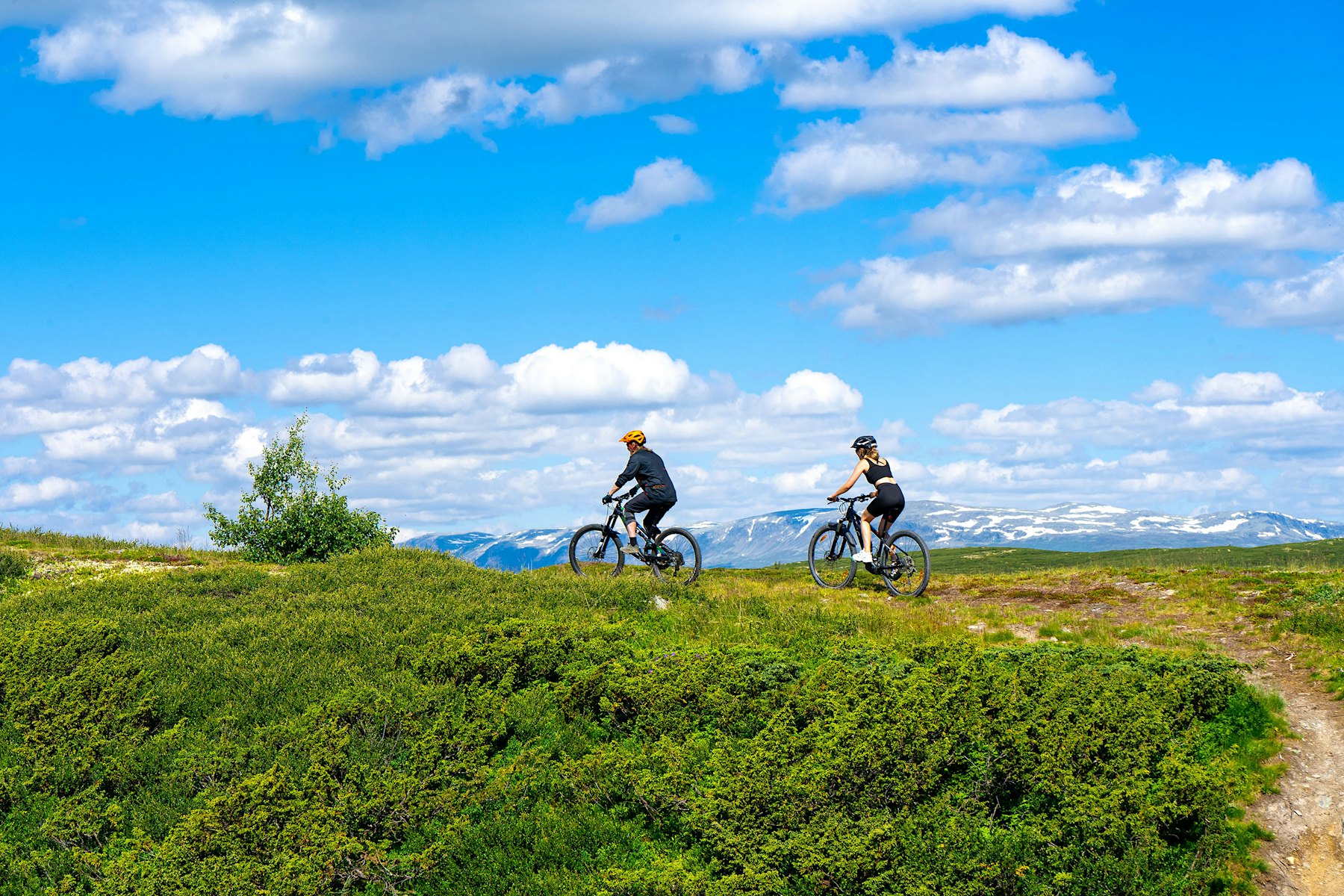 Two people are cycling on a path in the mountains, with several mountains in the background. Photo