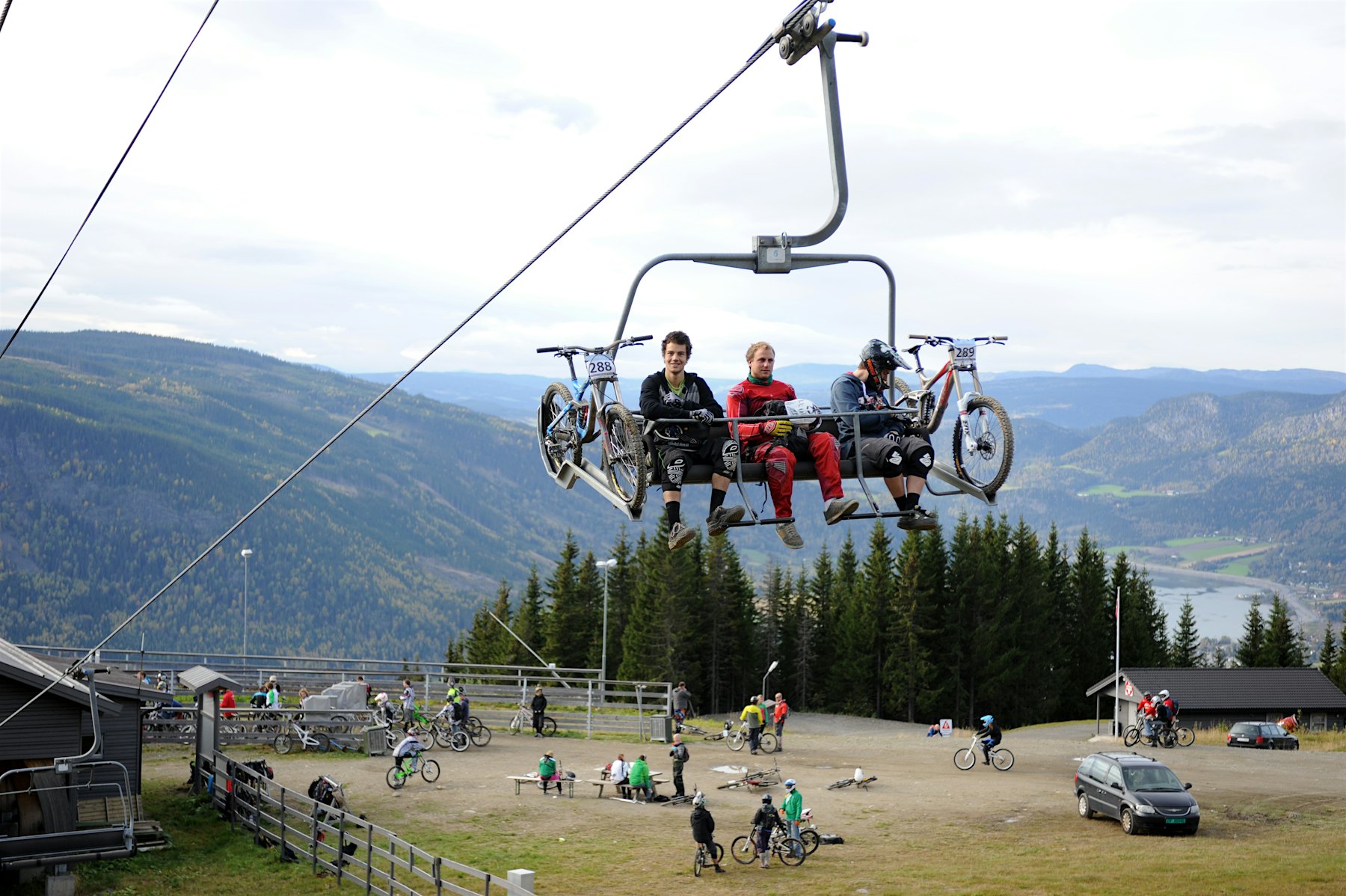 Three cyclists sit in a chairlift with their bikes, several cyclists on the ground below, mountains in the background. Photo