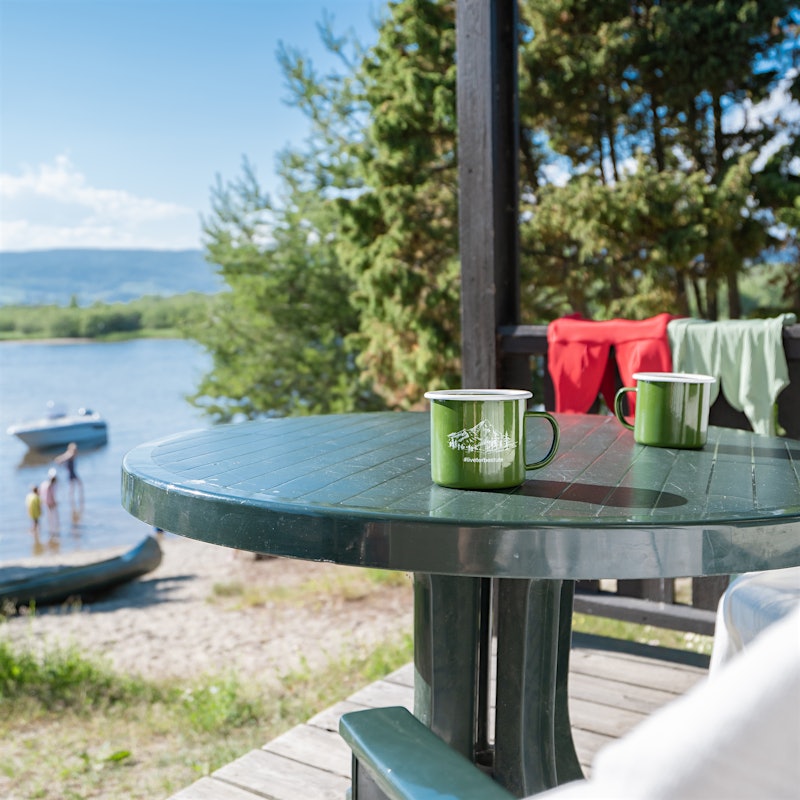 Terrasse med bord med to kopper på, to badedrakter henger på rekkverk i bakgrunnen, tre stykker bader i vannet nedenfor hytta, båter på vannet og kajakk på stranda.