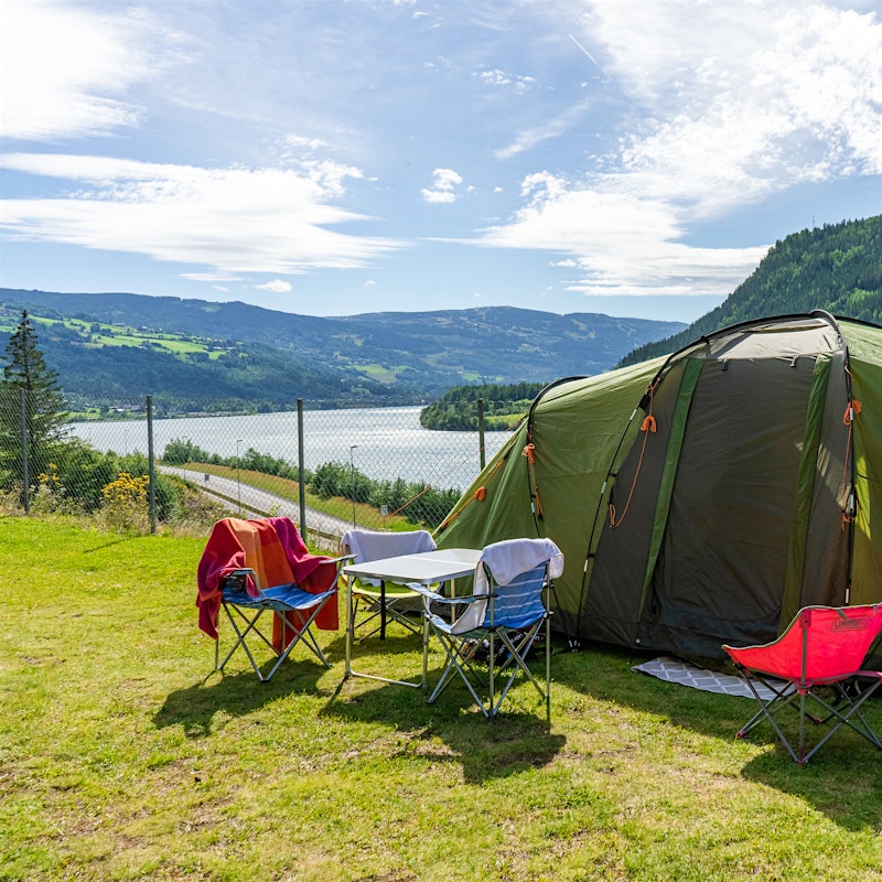 Telt står oppstilt på gress med utsikt over fjell og vann. Det er campingbord og stoler foran teltet.