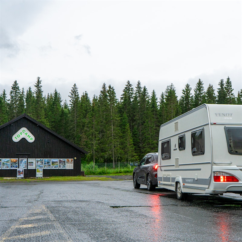 Bil med campingvogn står i innkjøring til campnigplass med resepsjonsbygget på siden.