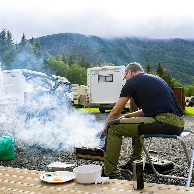 Man sits grilling in front of caravan. Mountains in the background. Photo