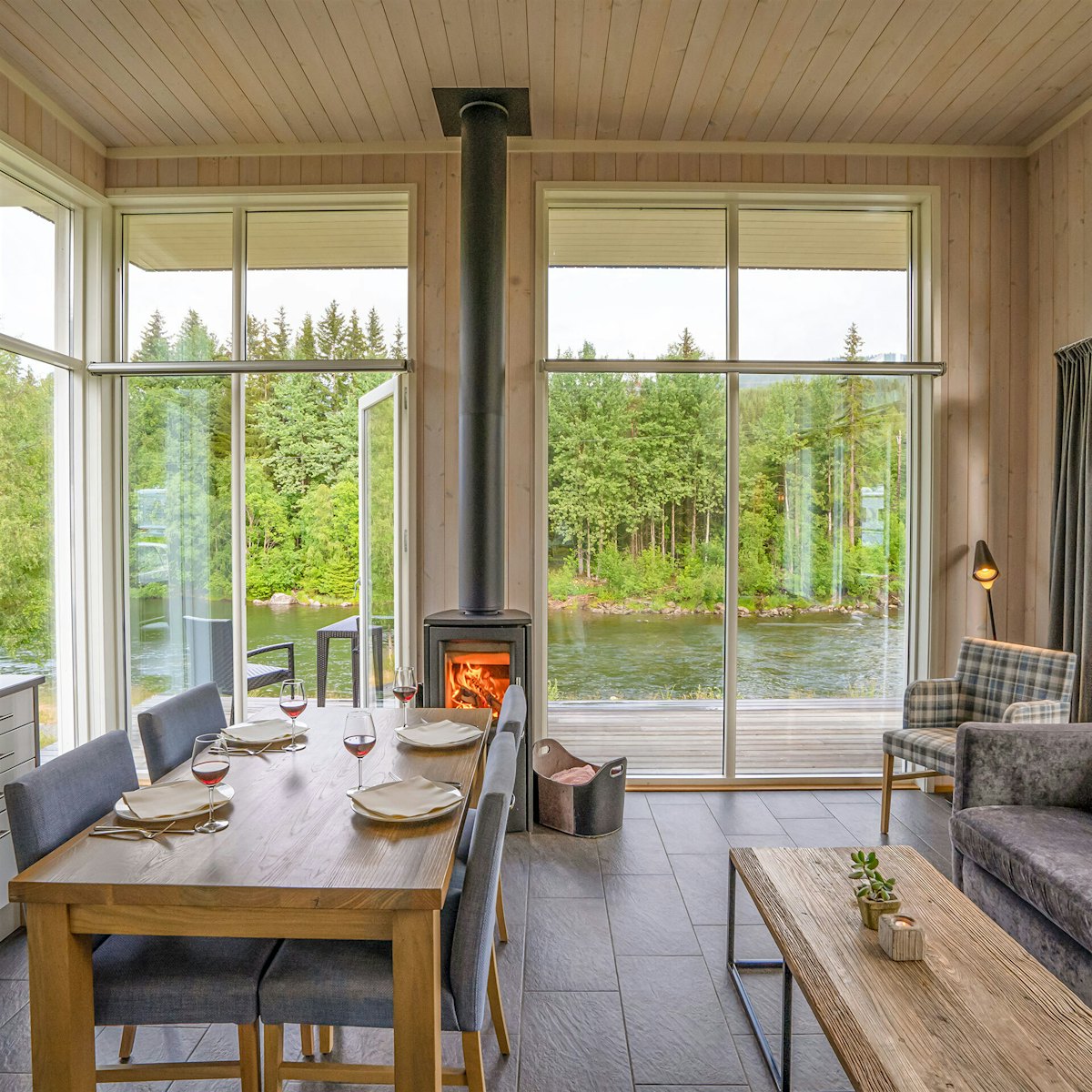 Lys stue med vinduer fra gulv til tak, spisebord sofa og peis. Utsikt over grønne trær og elv. Foto