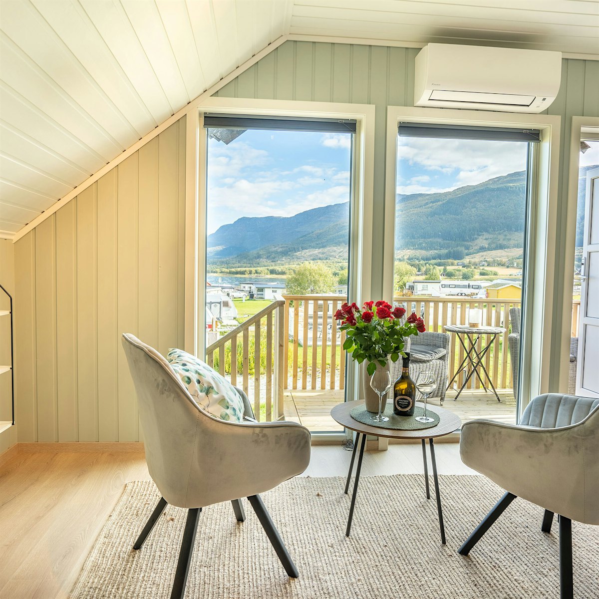 Helles und modernes Wohnzimmer mit großen Fenstern, zwei Stühlen und einem Tisch mit Rosen darauf. Blick auf Berge und Meer. Foto