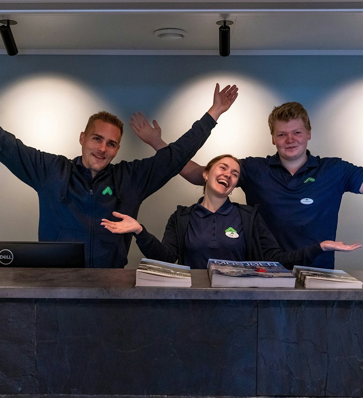 Three employees stand behind a reception and smile with raised hands.