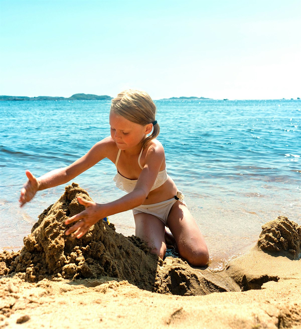 Jente sitter i strandkanten og bygger sandslott. Foto