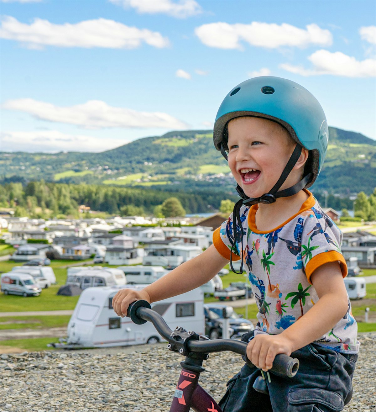 Boy smiles and laughs while sitting on a bicycle, with campsite in the background. Photo