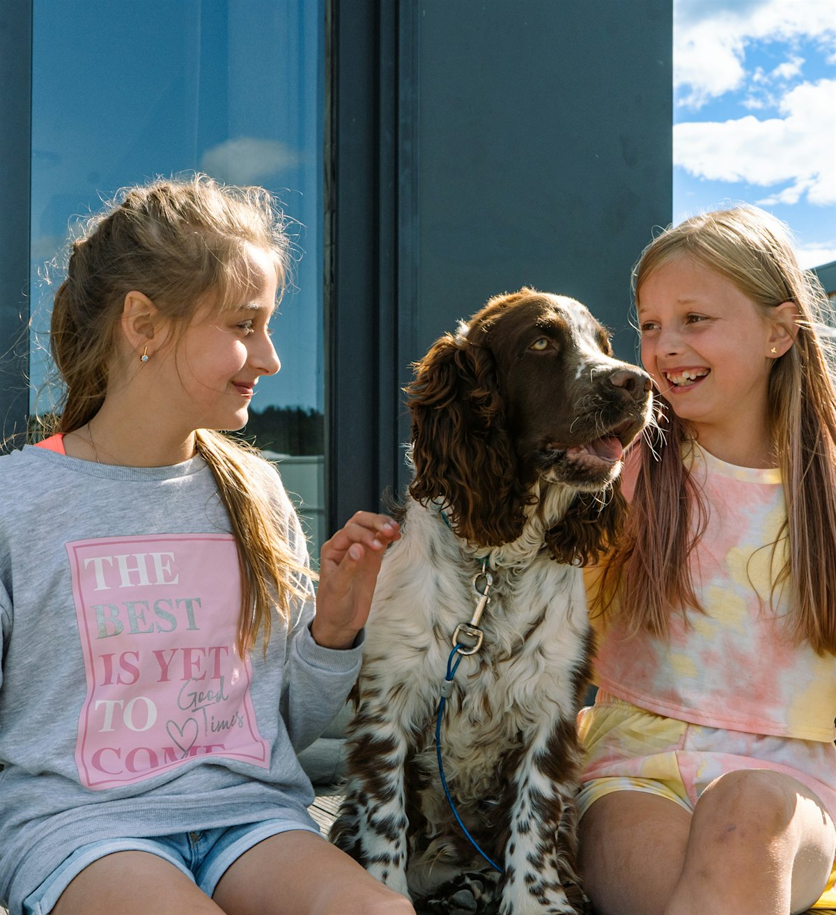 Two girls sit with a dog between them on a cottage terrace. Photo