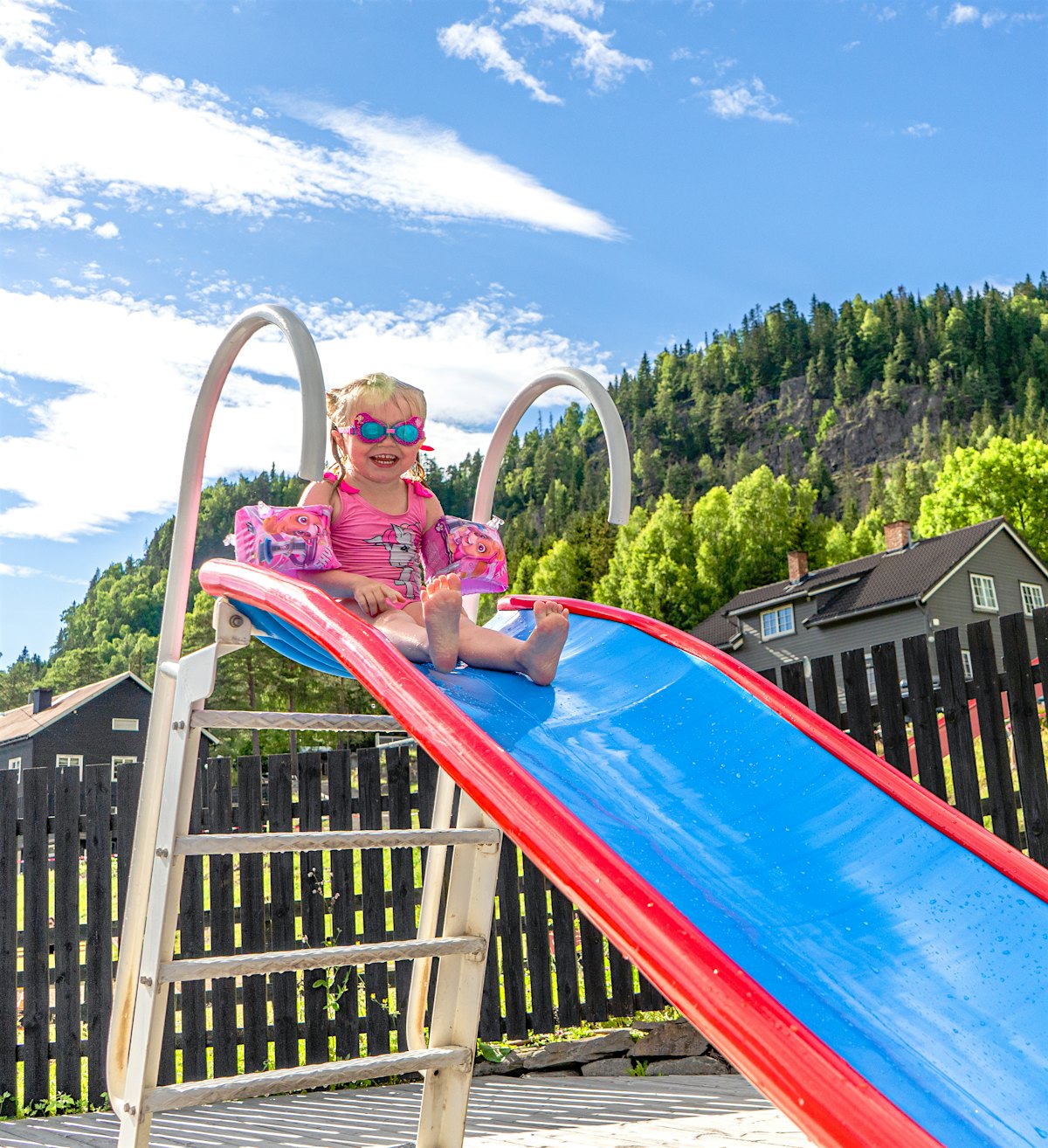 Girl sits on top of water slide and smiles, wearing bangles and swimming goggles. Photo