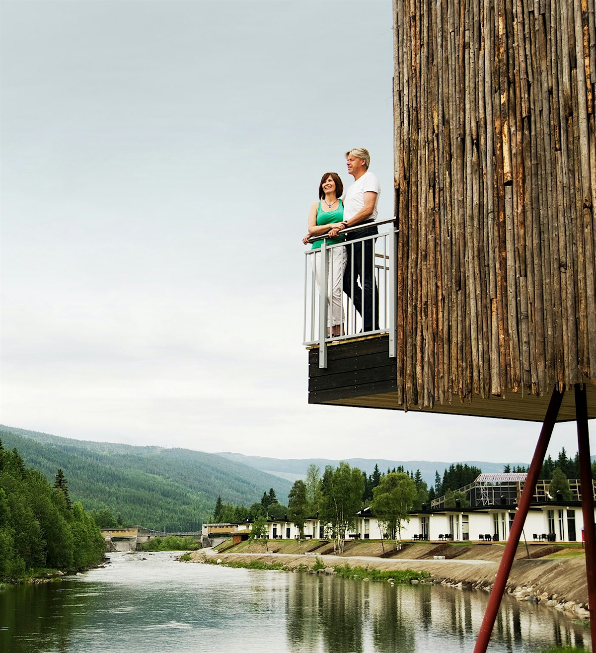 A couple stands together on the balcony of Styltehytta and looks out over the river and the forest. Photo
