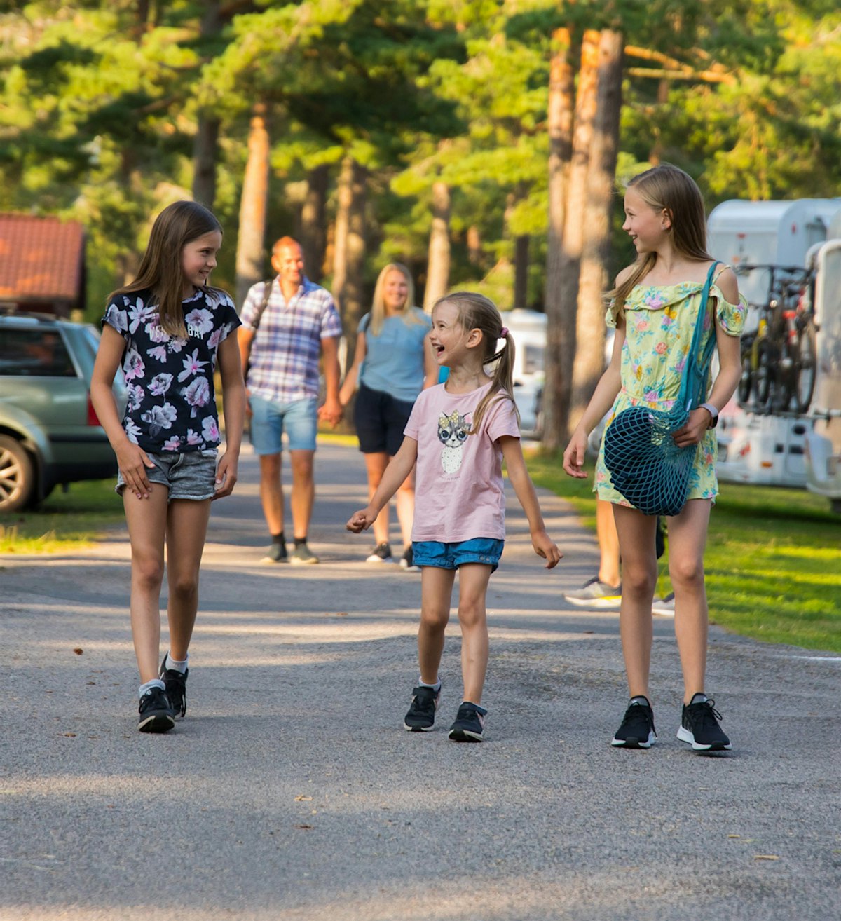 Drei Mädchen spazieren über den Campingplatz von Topcamp Sjøsanden und lächeln, ihre Eltern im Hintergrund. Foto
