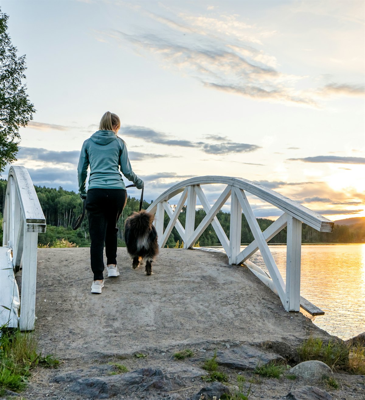 Kvinne går på tur med hund over bro i solnedgang. Foto