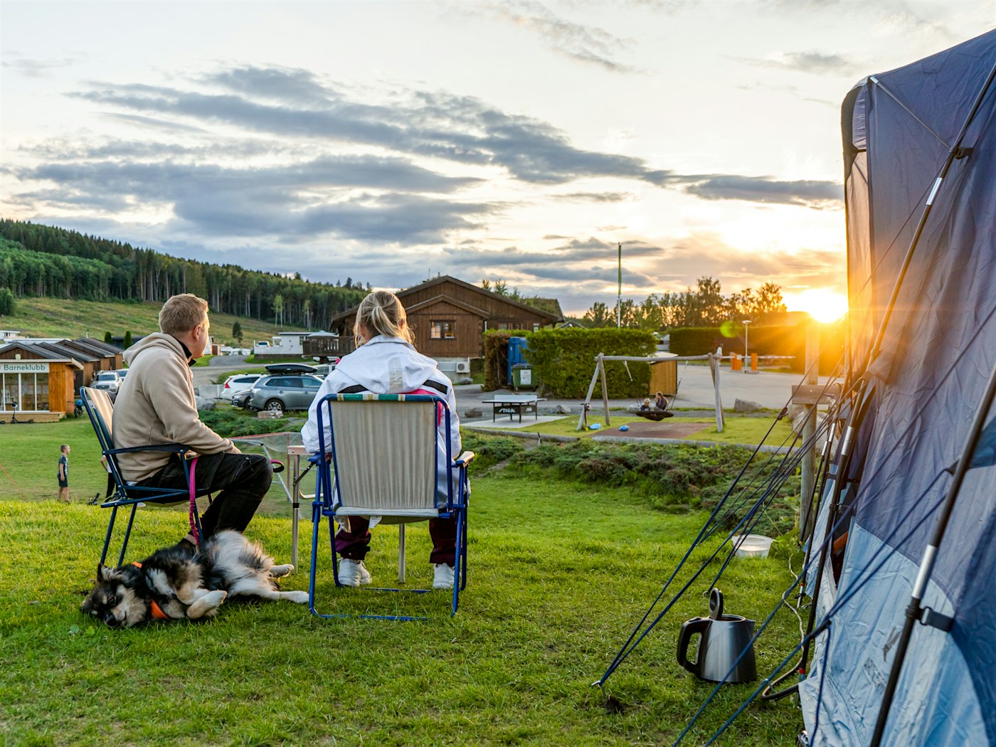 Mann, kvinne og hund sitter utenfor telt i solnedgangen. Foto